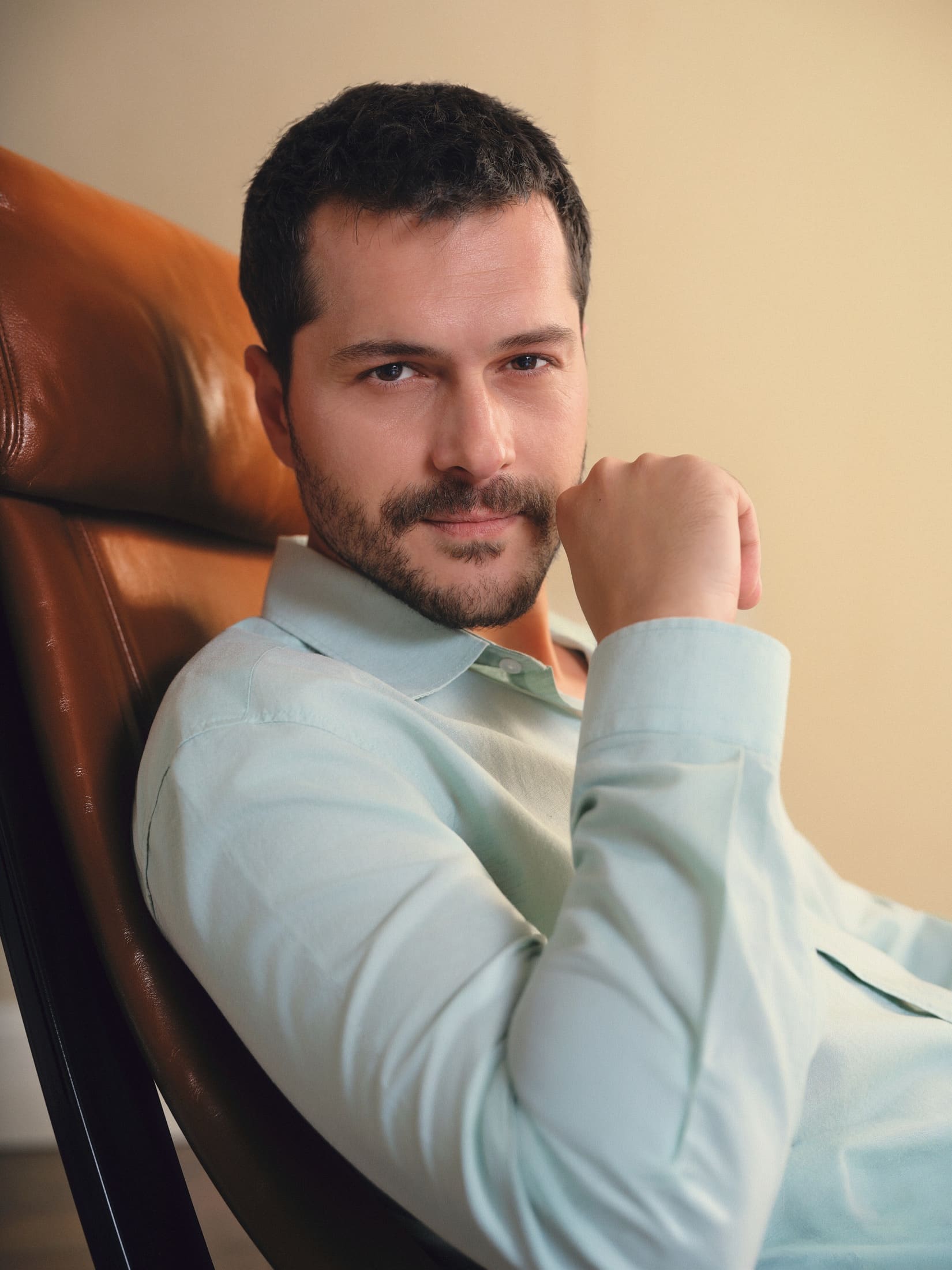 A close portrait of Alperen Duymaz reclining in a caramel leather lounge chair, fist to his chin in a pale mint shirt, for LC Waikiki.