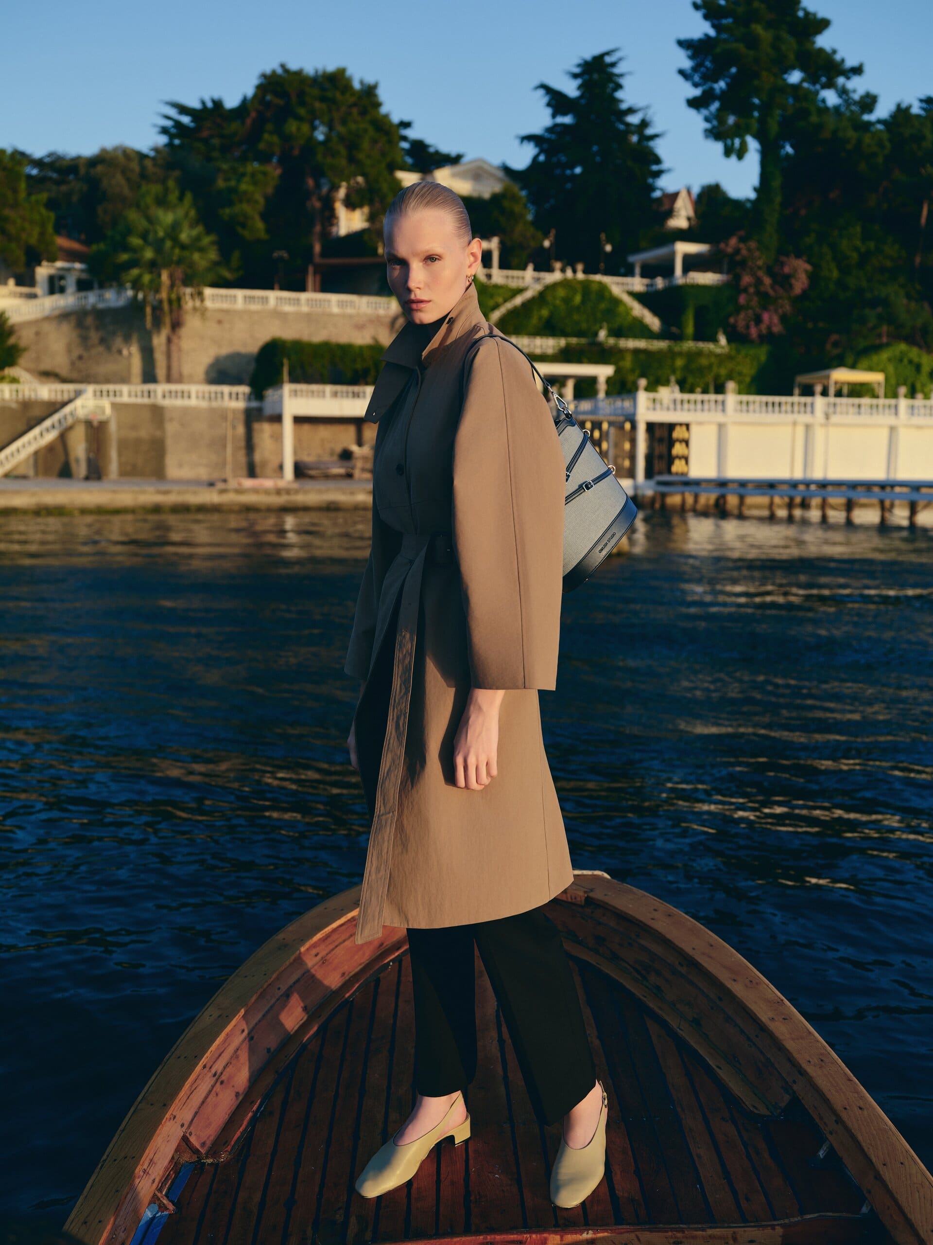 A belted taupe trench and two-tone canvas bucket bag carried on the prow of a wooden Bosphorus boat at golden hour for Owlish Studio.