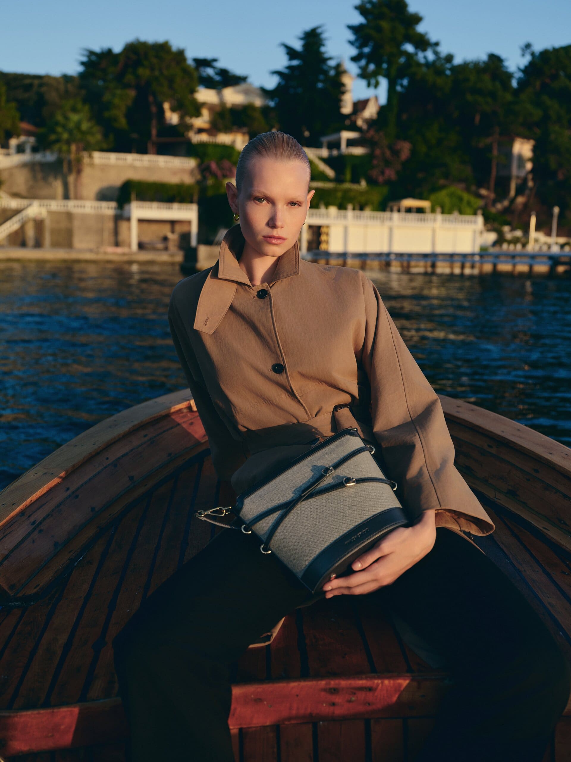 Perched on the bench of the wooden boat, the grey bucket bag squared on the knees against the shoreline mansions for Owlish Studio.