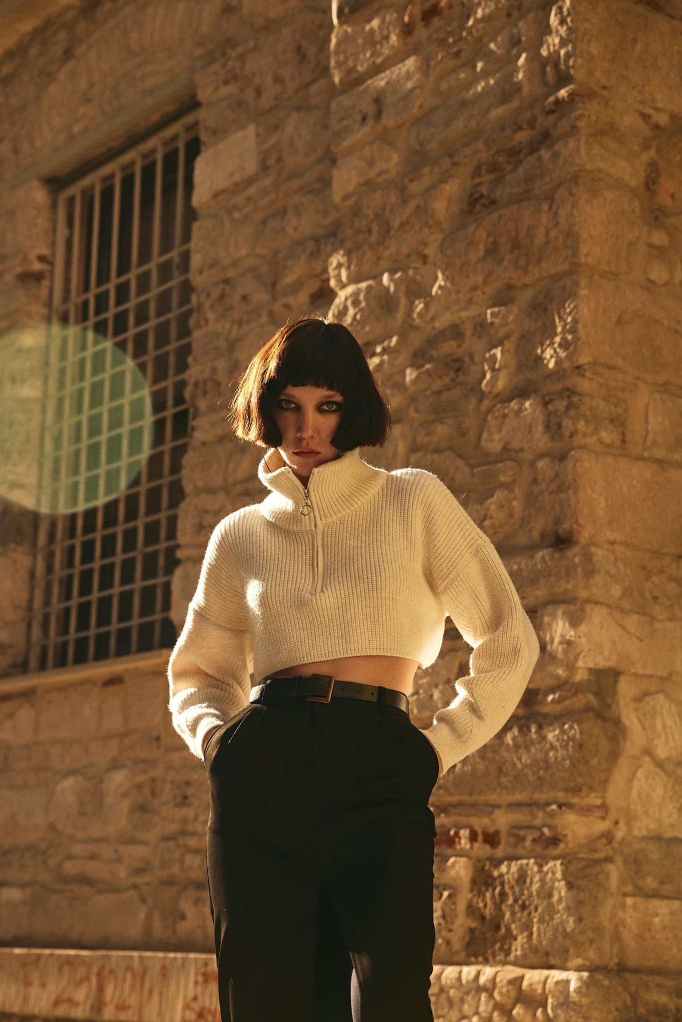 A cropped ivory zip-collar knit and belted black trousers in raking golden light against rough stone for Vatkalı.