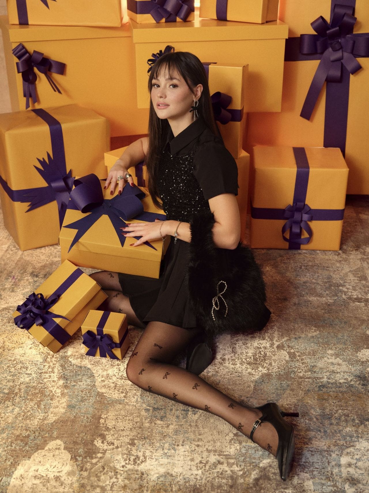 Derya Pınar Ak in a black sequin party dress with bow-print tights, seated among towering orange gift boxes tied in violet ribbon for XSIDE.