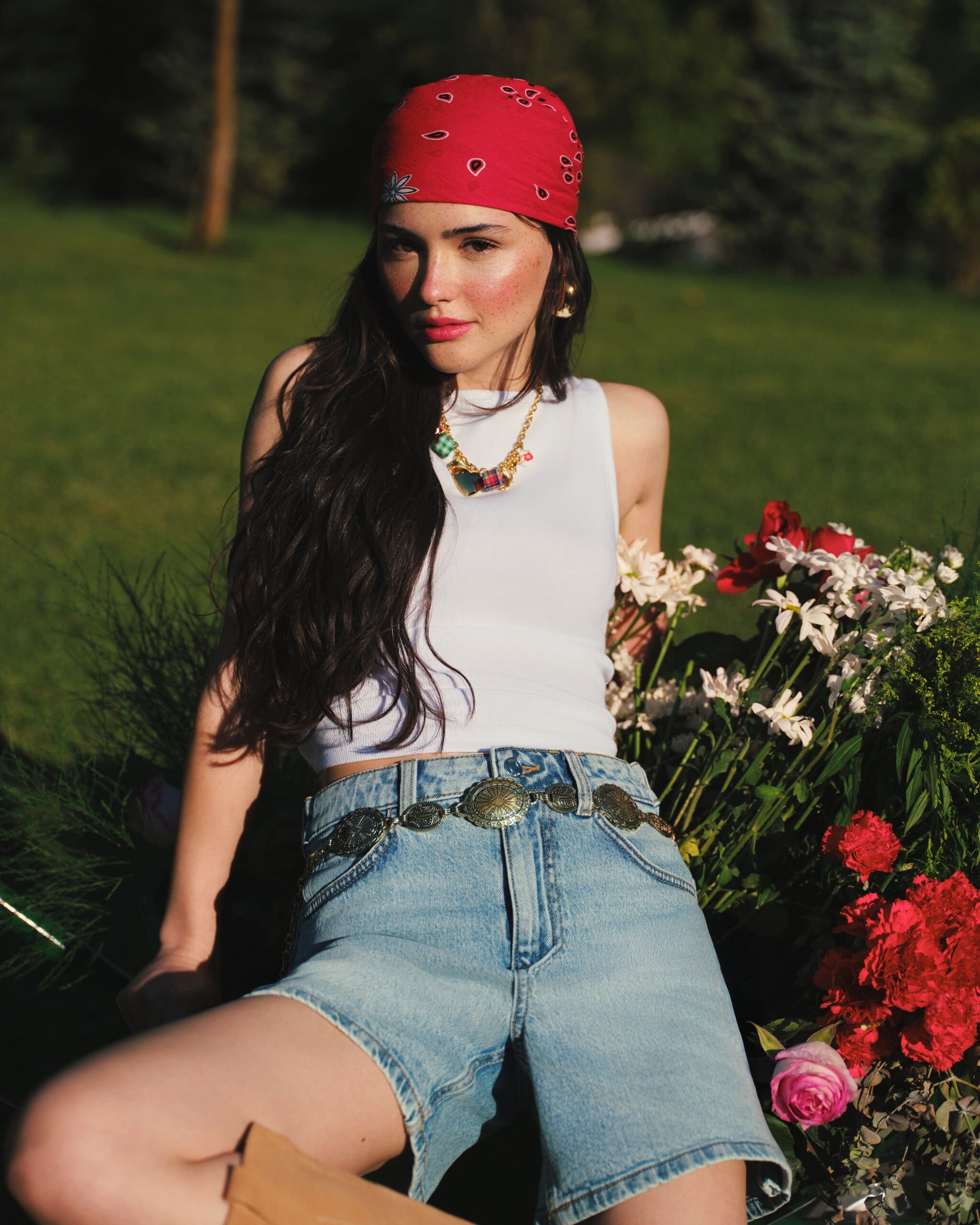 Derya Pınar Ak in a red paisley bandana and white tank, seated among carnations and daisies with a concho-belted denim short for XSIDE.