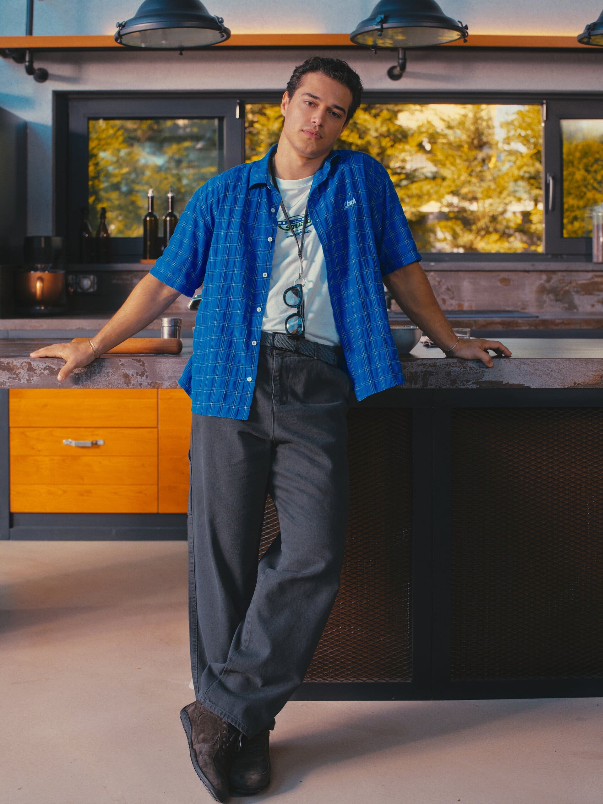 Kaan Miraç Sezen leans full-length against a steel kitchen counter in a cobalt check shirt for XSIDE.